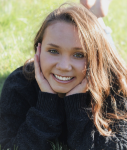 A woman laying in the grass, smiling with her hands cupping her cheeks. 